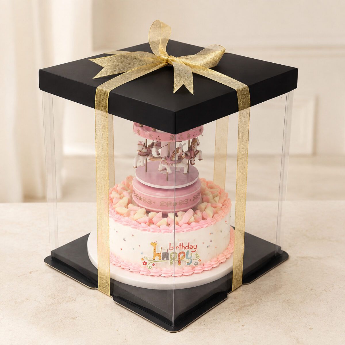 Tall clear square cake box with a black lid and base and a gold woven ribbon around a carousel cake