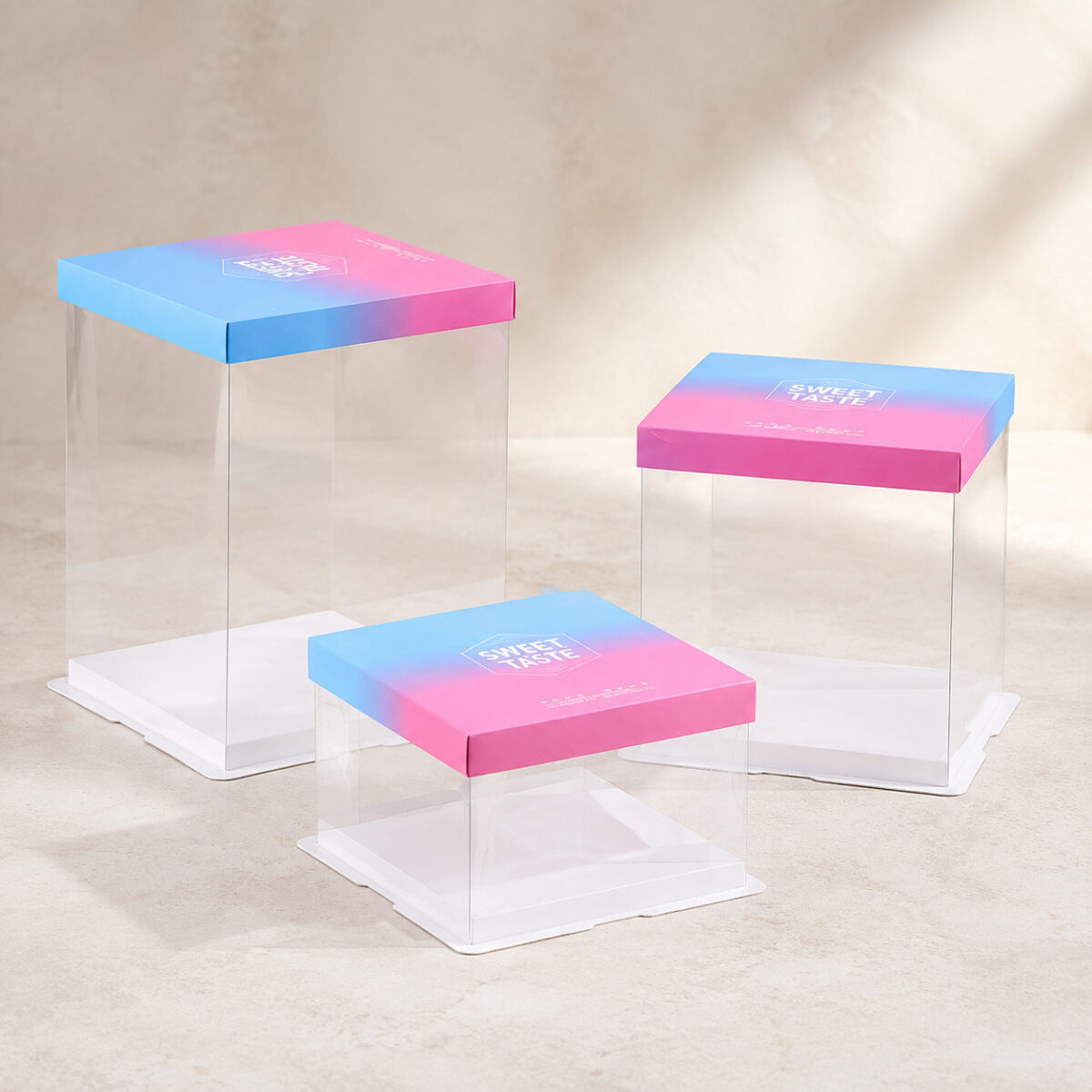 Three empty clear square cake boxes with blue and pink Sweet Taste lids and white bases