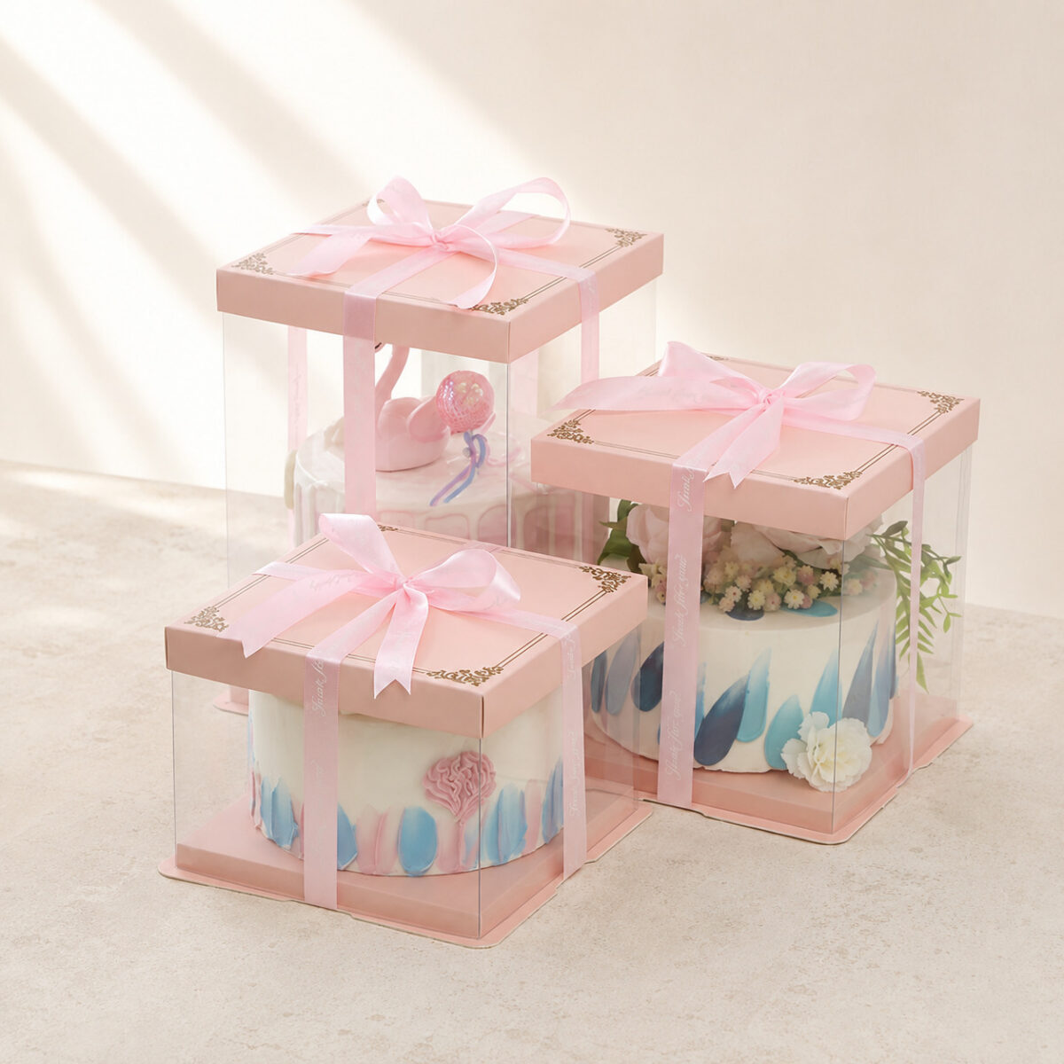 Three clear square cake boxes in different heights with pink lids, matching bases and pink ribbon bows