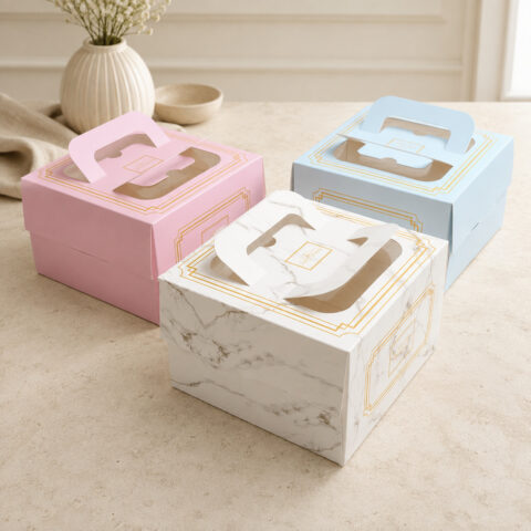 Three pink blue and marble-print square cake boxes with clear lid windows, folded paper handles and gold border artwork
