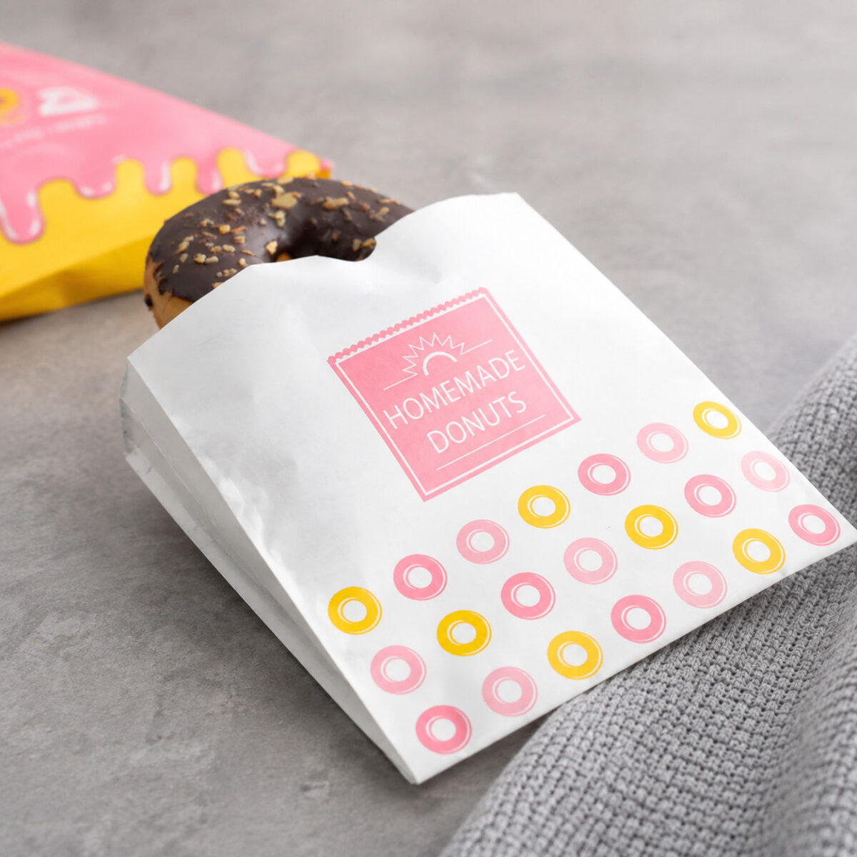 Printed Donut and Burger Paper Sleeves White printed donut paper sleeve holding a chocolate donut beside a pink and yellow package