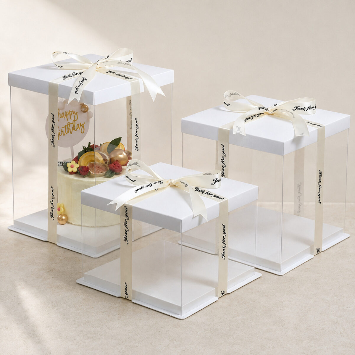 Three clear square cake boxes with white lids and ivory ribbons, shown in tall, medium and low heights