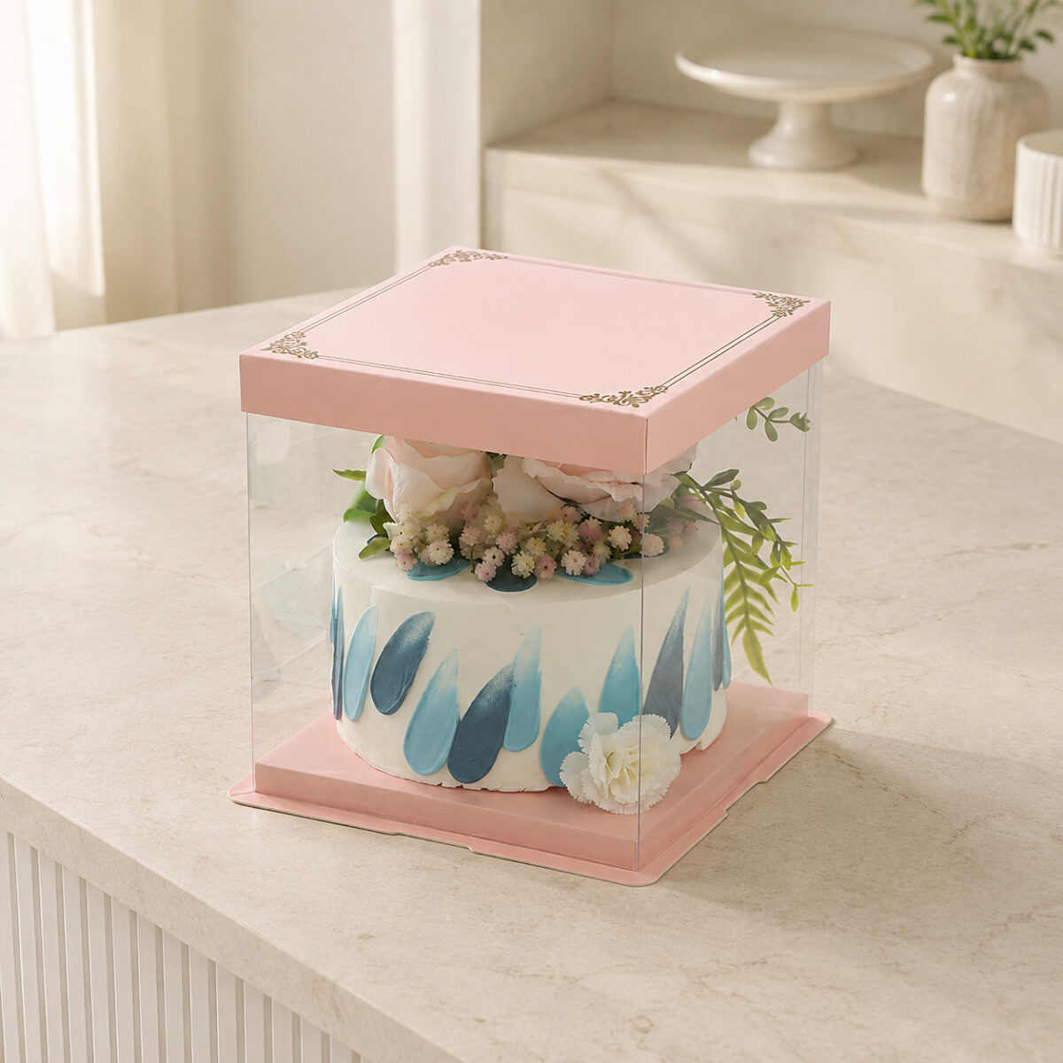 Clear square cake box with a blush pink lid and base enclosing a floral cake with blue decoration