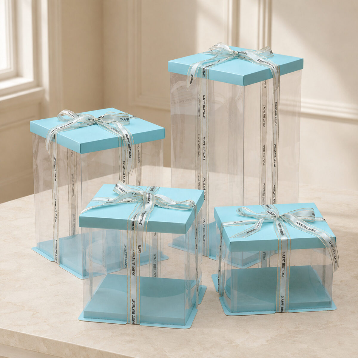 Four empty clear square cake boxes with blue lids and bases and Happy Birthday ribbons
