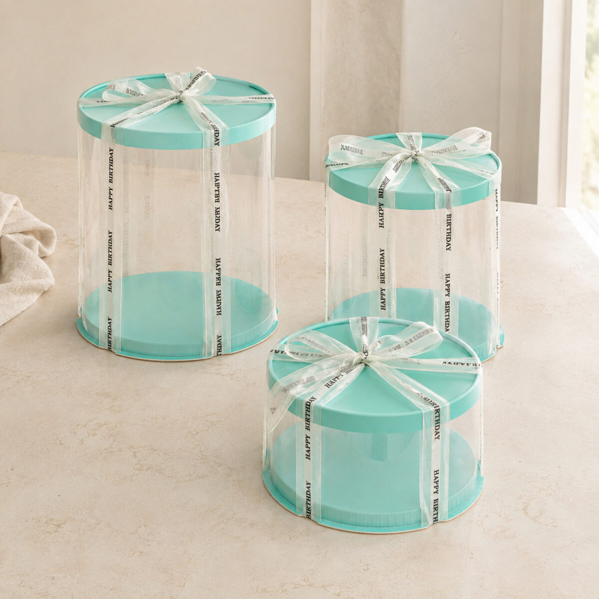 Three clear round cake boxes in tall medium and short heights with aqua lids and bases and ivory birthday ribbons