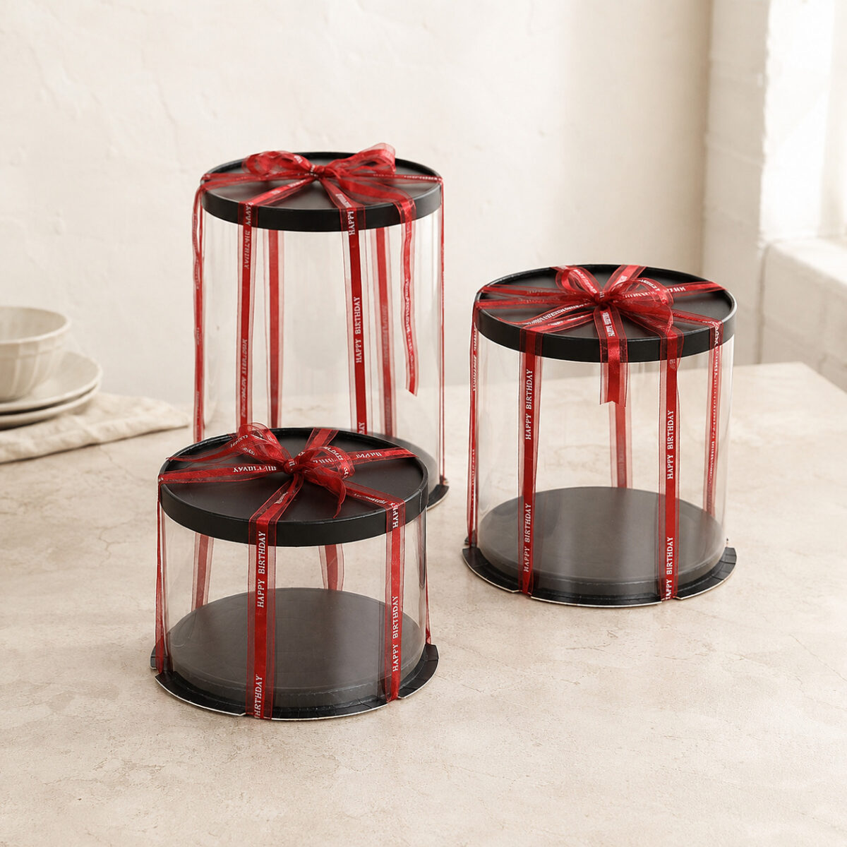 Three clear round cake boxes with black lids and bases and red Happy Birthday ribbons