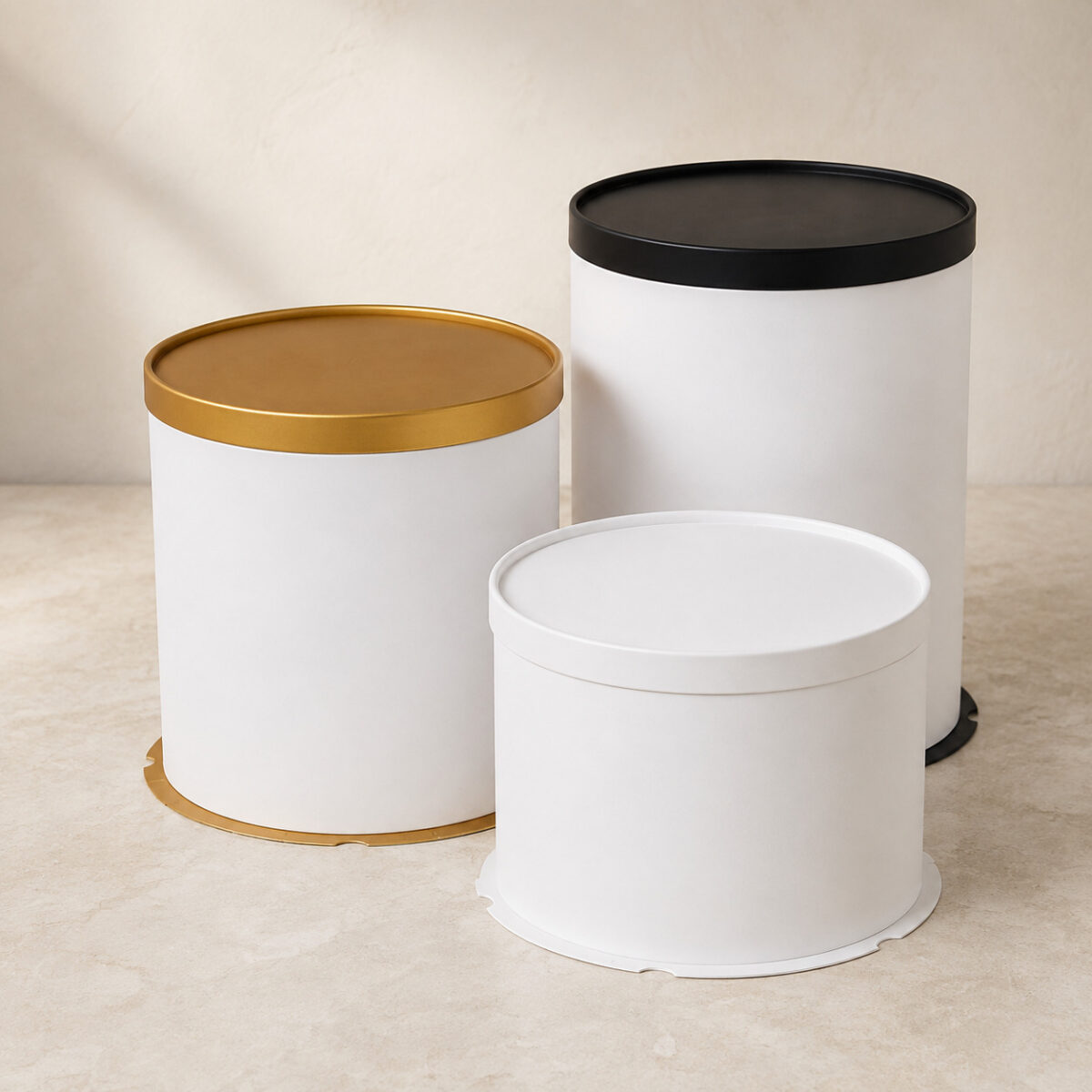 Three plain white cylindrical cake boxes with gold, black and white solid lids in different heights