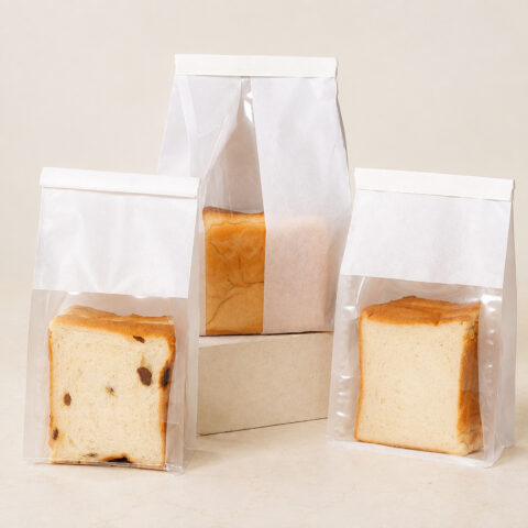 Three white paper bread bags with clear windows showing plain and raisin bread